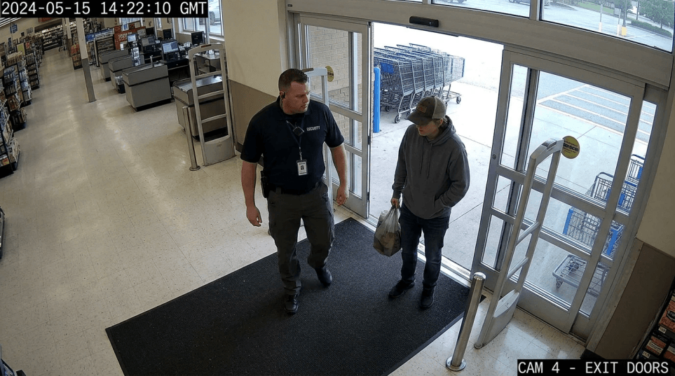 Overhead CCTV-style security camera view of a loss prevention agent approaching a person at the exit doors of a retail store, subject appears tense but stationary, realistic surveillance footage look