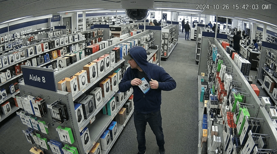 Overhead CCTV-style security camera footage perspective showing a person in a retail electronics store aisle slipping a small product into the inside pocket of their jacket, grainy realistic surveillance look, wide angle